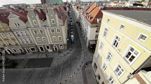 A drone flies over a narrow street in the old town of Lublin, Poland. The street is lined with historic buildings, many of which are decorated with colorful murals. There are a few cars parked on the
