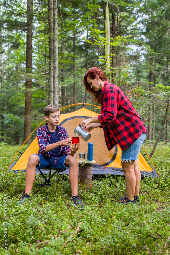 Fototapeta premium A family is relaxing at a campsite in the forest. Mother and son drink coffee near the tent.