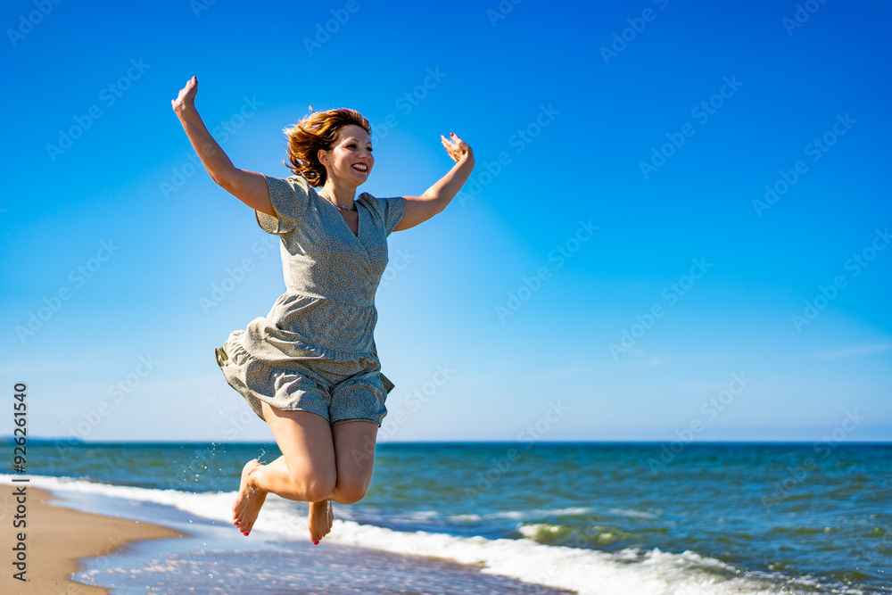 Young pretty woman wearing dress with raised arms jumping for joy high on beautiful sandy beach by sea shore on summer day. Power and joy of life Young pretty woman wearing dress with raised arms jumping for joy high on beautiful sandy beach by sea shore on summer day. Power and joy of life