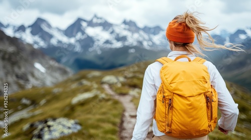 A woman with a bright orange backpack faces away from the camera, overlooking an expansive mountainous landscape. Her stance invites viewers to partake in the journey and exploration ahead.