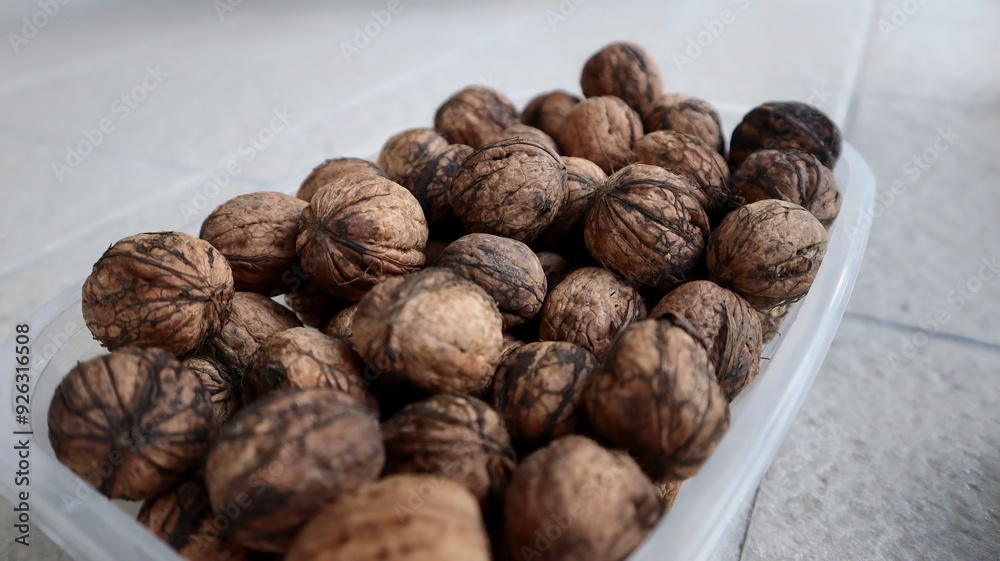 A close-up of a plastic container filled with whole walnuts. The walnuts have dark lines and strings (known as peduncles or fibers) on their shells, which are remnants from the husks. A close-up of a plastic container filled with whole walnuts. The walnuts have dark lines and strings (known as peduncles or fibers) on their shells, which are remnants from the husks.