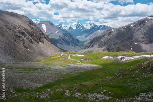 Mountain creek and lake against few big snowy pointy peaks far away. Sunlight and shadows of clouds in green alpine valley. Awesome scenic aerial view to three large beautiful snow-capped peaked tops.