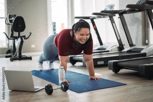 Smiling woman in a gym doing plank on a yoga mat, surrounded by treadmills, a laptop, water bottle, and weights.