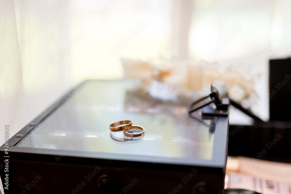 Wedding rings placed elegantly on a glass box near floral decor. Two wedding rings resting on a glass surface, surrounded by soft wedding decorations. Wedding rings placed elegantly on a glass box near floral decor. Two wedding rings resting on a glass surface, surrounded by soft wedding decorations.
