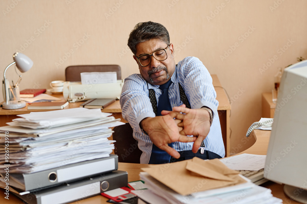 Obraz premium Stressed office worker stretching arms while sitting at cluttered desk with documents and files. Office environment showing piles of paperwork, computer, and traditional lamp