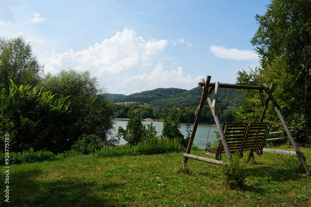 A wooden swing in a scenic Austrian countryside, surrounded by vibrant green grass, a clear blue sky, trees, and a calm river A wooden swing in a scenic Austrian countryside, surrounded by vibrant green grass, a clear blue sky, trees, and a calm river