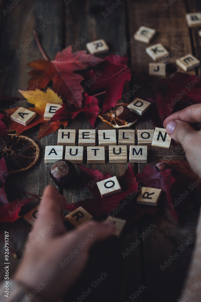 Obraz premium Woman's hands with Hello autumn text on dark wooden background. Composition with bright autumn leaves, pumpkin, dry citrus, walnut. Cozy fall mood concept. Flat lay, close up, top view, macro