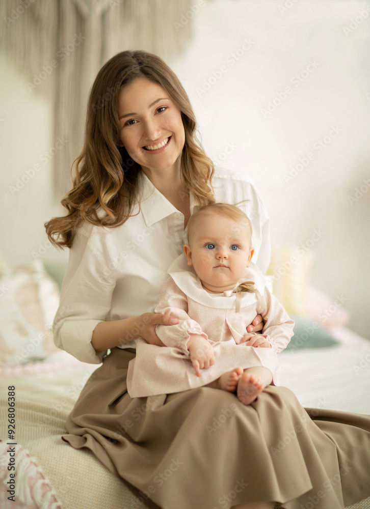 Beautiful young mother sits on the bed and holds her infant daughter on lap in delicate pastel colors interior. Maternal love and child care Beautiful young mother sits on the bed and holds her infant daughter on lap in delicate pastel colors interior. Maternal love and child care
