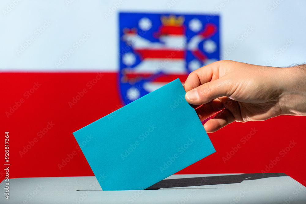 Voting envelope is put into ballot box for state election in Thuringia. Blue envelope in hand. Flag of Thuringia in the background. Election year 2024 in Germany. Voting envelope is put into ballot box for state election in Thuringia. Blue envelope in hand. Flag of Thuringia in the background. Election year 2024 in Germany.