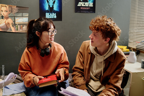 Two students sitting in cozy bedroom setting, discussing study material and holding books. Anime posters on walls and comfortable atmosphere indicating relaxed study environment