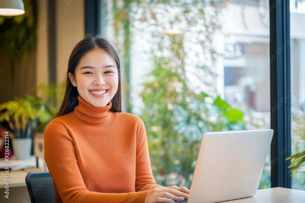 Young Asian woman smiling while working on a laptop in a modern office with large windows and natural light, surrounded by green plants and bright lighting, creating a warm and welcoming atmosphere.