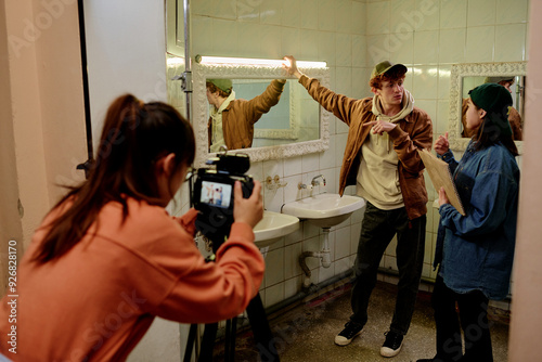 Young man pointing while being filmed by photographer in bathroom scene setting Photographers capturing moments in dramatic performance setting with detailed backdrop photo