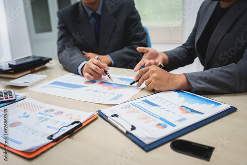 Asian business team engages in a collaborative meeting, financial charts at a desk. Dressed in formal suits, they brainstorm creative ideas to enhance company performance and team cohesion.