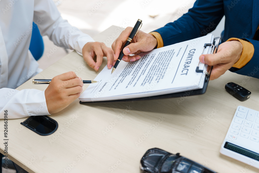 Fototapeta premium A middle-aged Asian male seller in a formal suit assists a woman buyer at a desk. They finalize a vehicle lease, reviewing financial charts and signing contract agreements with a car key.