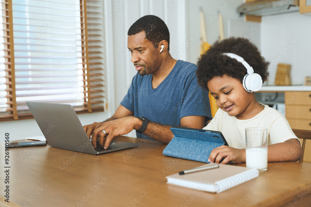 Black African American Father and child using laptop and tablet with headphones at home kitchen table. Casual home lifestyle. Family bonding and home learning concept. Black African American Father and child using laptop and tablet with headphones at home kitchen table. Casual home lifestyle. Family bonding and home learning concept.