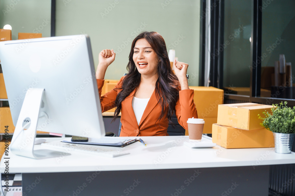 A beautiful businesswoman works at her desk, selling products online. She smiles as she looks at the computer screen, satisfied that her online business has exceeded sales targets.