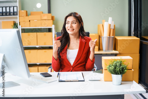 A beautiful businesswoman sits at the table, looking at the computer screen and talking on the phone. She analyzes marketing plans with a smile, feeling happy and successful in her online business.