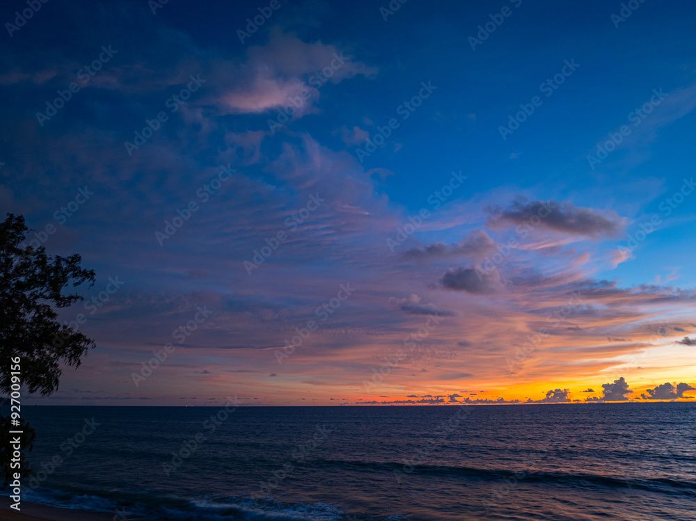 Obraz premium A captivating scene of a vibrant sunset casting an orange and purple glow across the sky, with gentle ocean waves lapping at the shoreline. The dramatic cloud formations add depth to the serene.