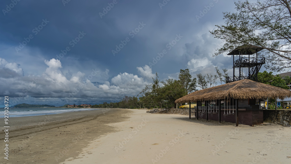 Fototapeta premium A bar on a sandy beach. Thatched roof. Swing seats are suspended at the bar counter. A lifeguard observation tower against a blue sky, clouds. The waves of the turquoise ocean are foaming. Malaysia
