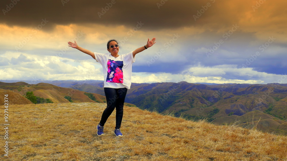 junge sportliche Frau auf einem Berggipfel in Sumba streckt die Arme hinauf zu dramatischen Wolken junge sportliche Frau auf einem Berggipfel in Sumba streckt die Arme hinauf zu dramatischen Wolken