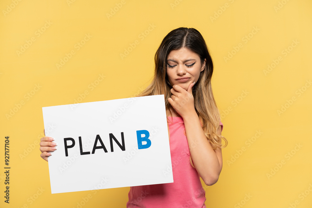 Young Russian girl isolated on yellow background holding a placard with the message PLAN B and thinking Young Russian girl isolated on yellow background holding a placard with the message PLAN B and thinking