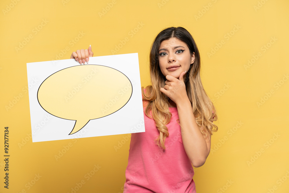 Young Russian girl isolated on yellow background holding a placard with speech bubble icon and thinking Young Russian girl isolated on yellow background holding a placard with speech bubble icon and thinking