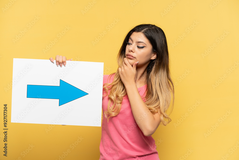 Young Russian girl isolated on yellow background holding a placard with arrow symbol and thinking