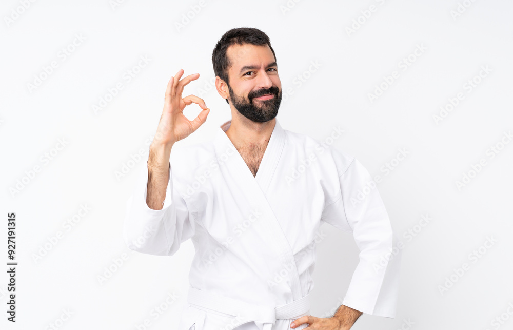Young man doing karate over isolated white background showing ok sign with fingers Young man doing karate over isolated white background showing ok sign with fingers