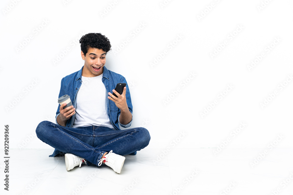 Venezuelan man sitting on the floor holding coffee to take away and a mobile Venezuelan man sitting on the floor holding coffee to take away and a mobile