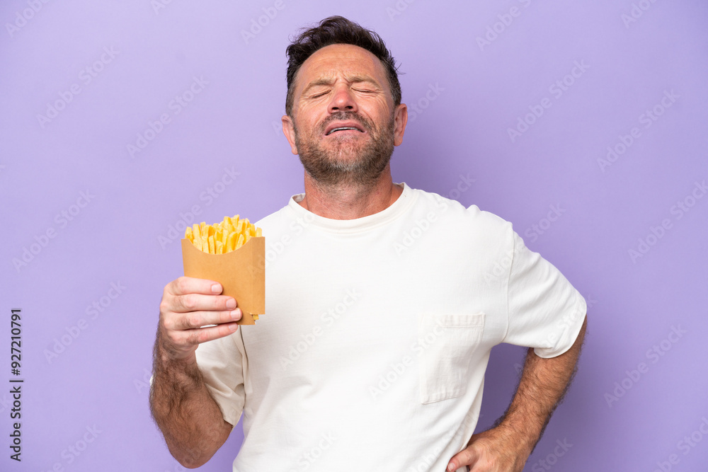 Middle age caucasian man holding fried chips isolated on purple bakcground suffering from backache for having made an effort Middle age caucasian man holding fried chips isolated on purple bakcground suffering from backache for having made an effort