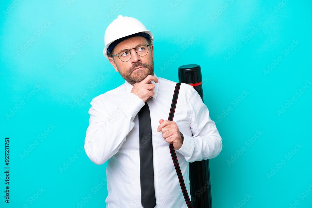 Middle age architect man with helmet and holding blueprints isolated on blue background and looking up Middle age architect man with helmet and holding blueprints isolated on blue background and looking up