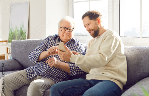 Happy adult son helps elderly father understand the phone and modern technology, sitting together on the sofa at home. Child learning old parent about the internet and online tools.