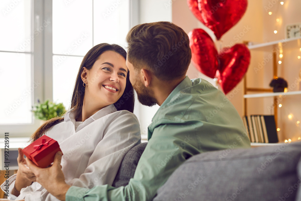 Young couple in love sitting on the sofa at home and exchanging romantic gifts. Man gives a present to a happy, smiling beautiful young woman on St Valentine's Day. Love and romance concept Young couple in love sitting on the sofa at home and exchanging romantic gifts. Man gives a present to a happy, smiling beautiful young woman on St Valentine's Day. Love and romance concept