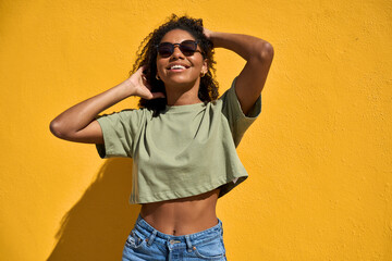 Cheerful cool young Black woman wearing brown sunglasses standing at yellow wall background. Happy smiling African American gen z ethnic girl feeling joy enjoying sunny summer day outdoors.
