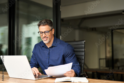 Happy busy older professional businessman entrepreneur using laptop holding bill corporate document in office. 50 years old man executive manager or business owner reading paper working on computer photo