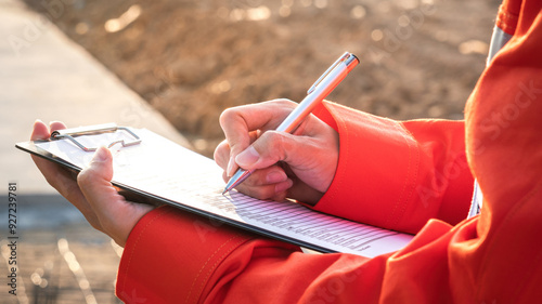 A project engineer in orange coverall uniform is using a pen to checking on building quality checklist to inspecting the quality, with background of construction work site. Close-up. photo