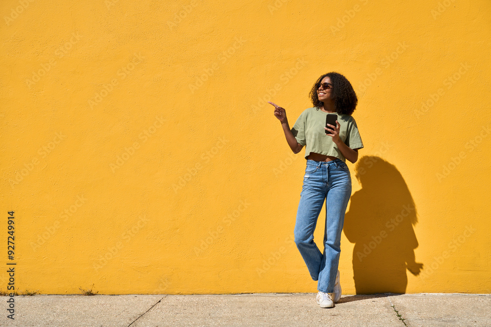 Obraz premium Happy African American gen z girl wearing sunglasses standing at yellow wall pointing at copy space holding smartphone. Smiling young Black woman using mobile phone advertising summer sale. Full body.