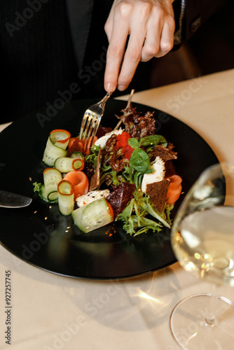 handsome man in a black sweater is eating a delicious salad with juicy meat, fresh vegetables, fragrant herbs, and crispy croutons, served on a black plate in a cozy, charming restaurant