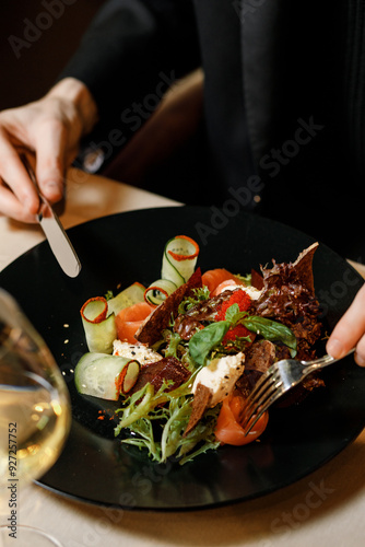 handsome man in a black sweater is eating a delicious salad with juicy meat, fresh vegetables, fragrant herbs, and crispy croutons, served on a black plate in a cozy, charming restaurant