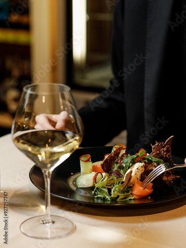 handsome man in a black sweater is eating a delicious salad with juicy meat, fresh vegetables, fragrant herbs, and crispy croutons, served on a black plate in a cozy, charming restaurant