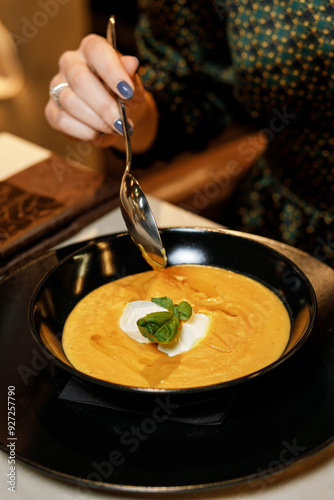 cute girl in a beautiful dress is eating a delicious cream soup with cream cheese, fresh vegetables, and fragrant herbs, served on a white plate in a cozy, charming restaurant