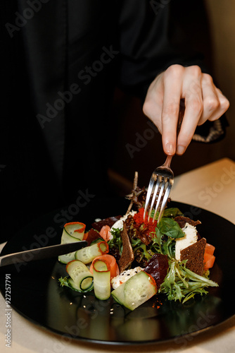 handsome man in a black sweater is eating a delicious salad with juicy meat, fresh vegetables, fragrant herbs, and crispy croutons, served on a black plate in a cozy, charming restaurant