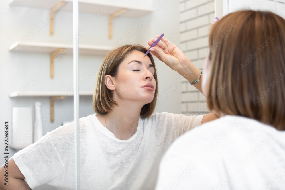 Young woman depilates her eyebrows and gives them the right shape with eyebrow tweezers in front of the mirror. Beauty and self care concept