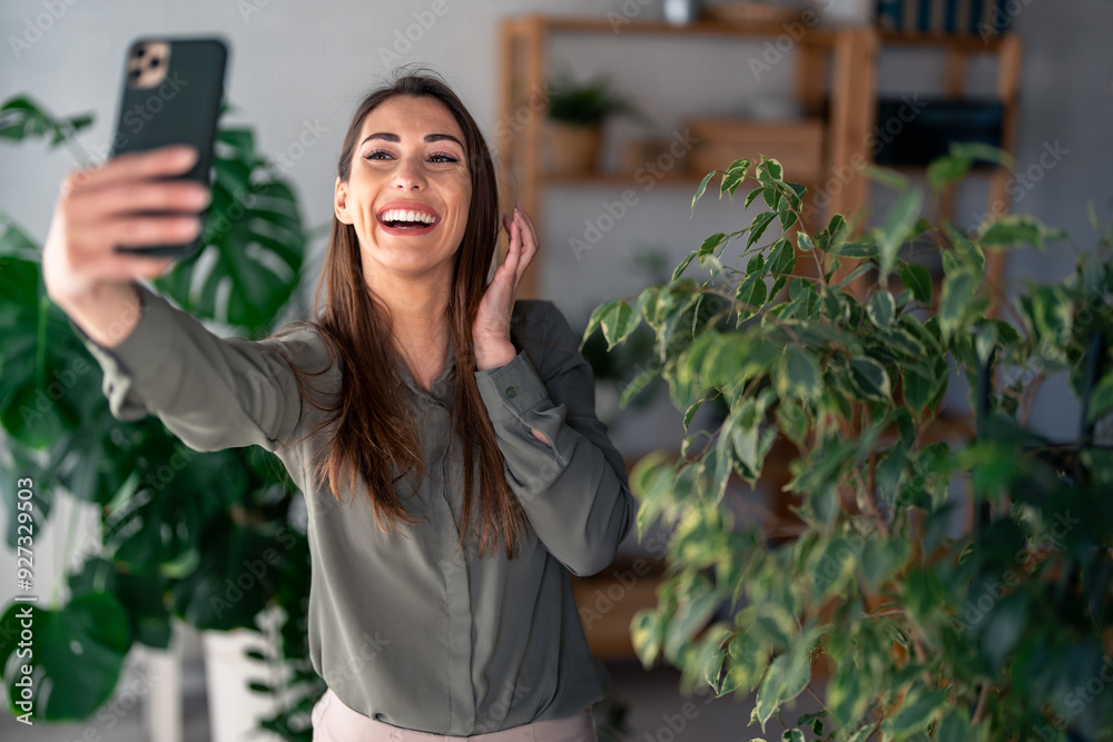 A cheerful caucasian lady engages in taking a selfie with her smartphone, clad in casual elegant attire, surrounded by trendy home decor and green plants. A cheerful caucasian lady engages in taking a selfie with her smartphone, clad in casual elegant attire, surrounded by trendy home decor and green plants.