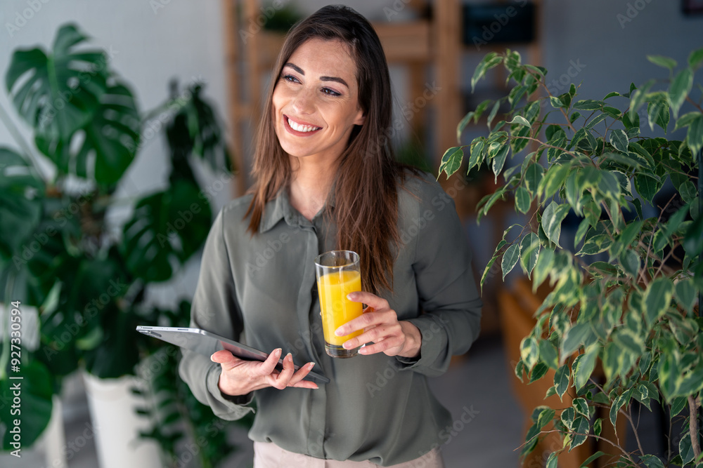 A cheerful Caucasian woman enjoys a fresh glass of juice while expertly navigating a digital tablet in her cozy home office, surrounded by lush greenery.