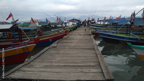 A scenic view of a wooden dock leading to colorful fishing boats adorned with Indonesian flags. The tranquil harbor scene features calm waters and distant mountains under a cloudy sky. photo