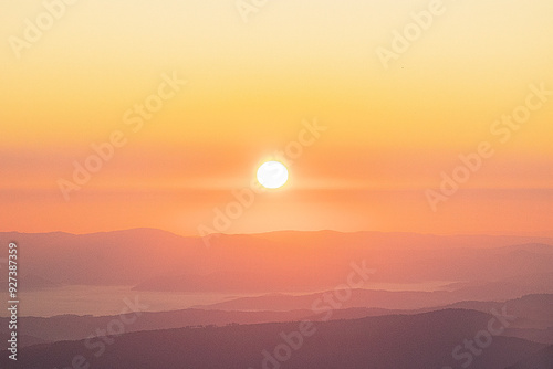 Bright sun rising over hazy mountain ranges, casting warm yellow and orange light across the landscape, creating a soft and serene morning atmosphere