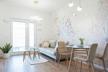 Modern living room with dining table with stylish chairs. White wall with wallpaper and luxurious interior in apartment with window. Hotel with furniture. Real estate.	