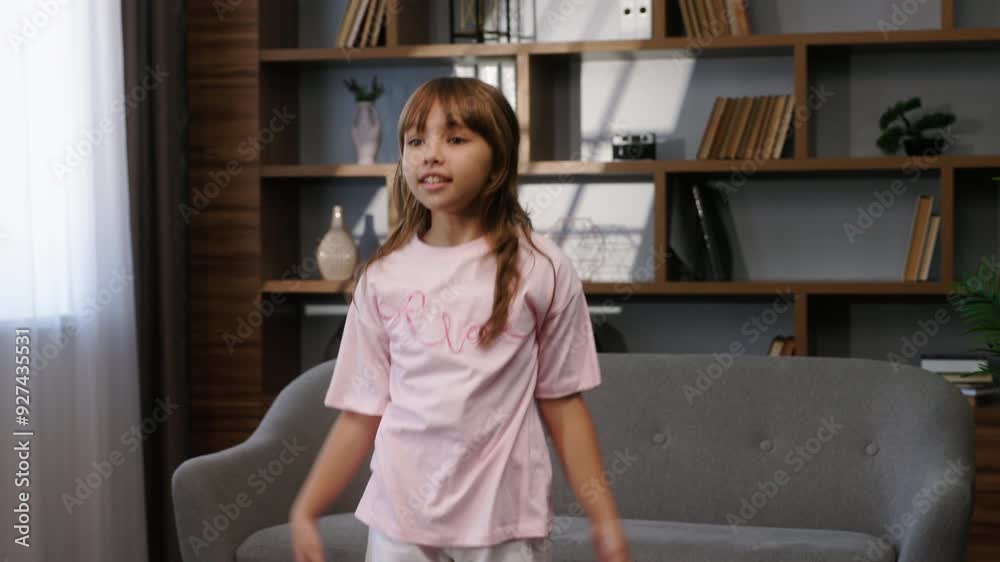 A little beautiful girl is exercising at home on a mat. The child is smiling and jumping while doing jumping jacks. In the background, there is a couch and a bookshelf. medium shot A little beautiful girl is exercising at home on a mat. The child is smiling and jumping while doing jumping jacks. In the background, there is a couch and a bookshelf. medium shot