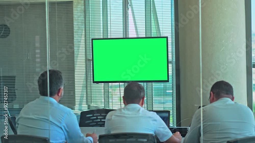 Green screen monitor in a conference room with large windows. View from the back through the glass walls on a company meeting during the day. Blinds on a transparent glass wall